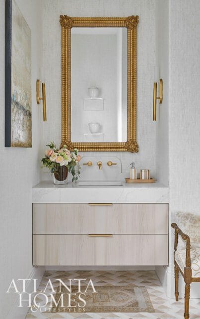 Powder room with a marble countertop, light wood vanity with brass hardware, ornate gold mirror, and textured wallpaper.