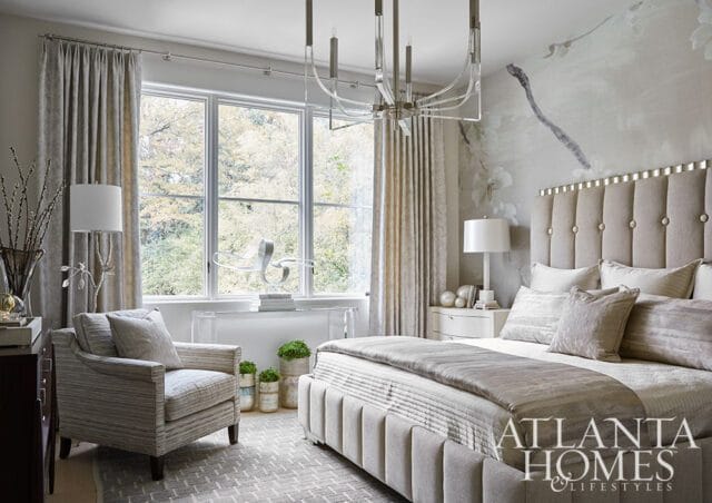 Neutral bedroom with tufted headboard, upholstered bed, armchair, and large window overlooking trees. Atlanta Homes & Lifestyles logo visible.