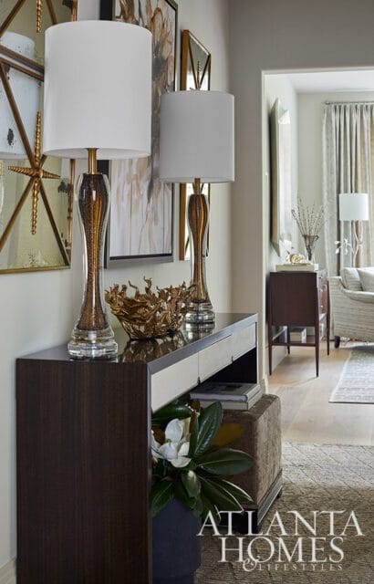 Dark wood console table with white and cream drawers, topped with two ornate glass lamps, a gold sculptural bowl, and artwork. A magnolia branch is in a dark vase at the base.