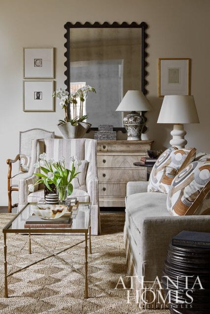 Living room with striped armchair, glass coffee table with tulips, and a sofa with patterned pillows. A large mirror hangs above a wooden dresser.