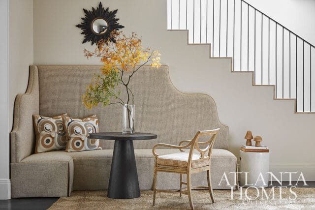 Upholstered banquette seating with patterned pillows, a dark pedestal table with a vase of fall branches, and a wooden chair next to a staircase with black railings.