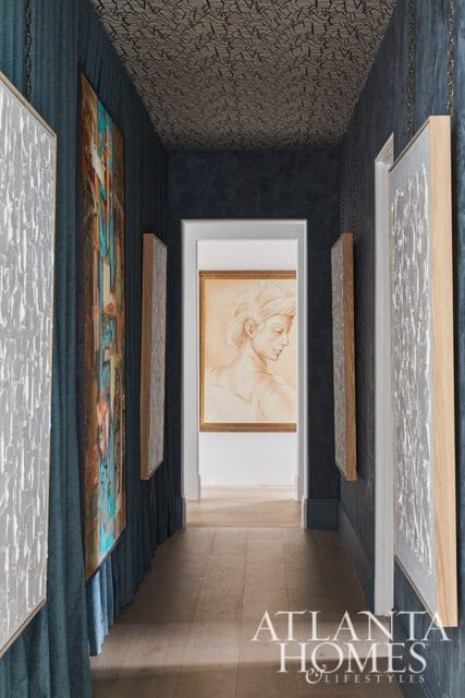 Hallway with dark blue textured wallpaper, patterned ceiling, and wood floors. Artwork hangs on the walls, including a framed sketch visible through an open doorway.