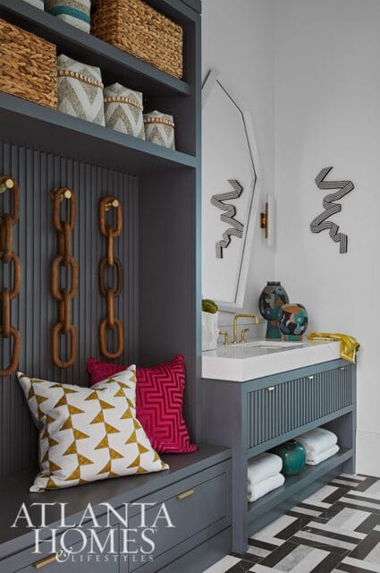 Gray built-in cabinetry with woven baskets on shelves, decorative chain hooks, and geometric pillows on a bench next to a vanity with a white countertop, brass faucet, and fluted cabinet doors.