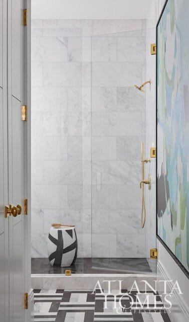 Modern bathroom with marble tile shower, brass fixtures, graphic stool, and patterned floor.