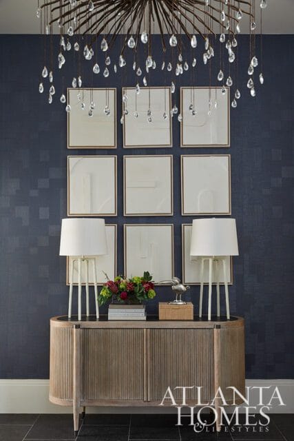 Dark blue textured wallpaper adorns the wall behind a wooden credenza topped with two white lamps, a floral arrangement, and a silver bird sculpture. Nine framed abstract artworks hang above the credenza, beneath a crystal chandelier.
