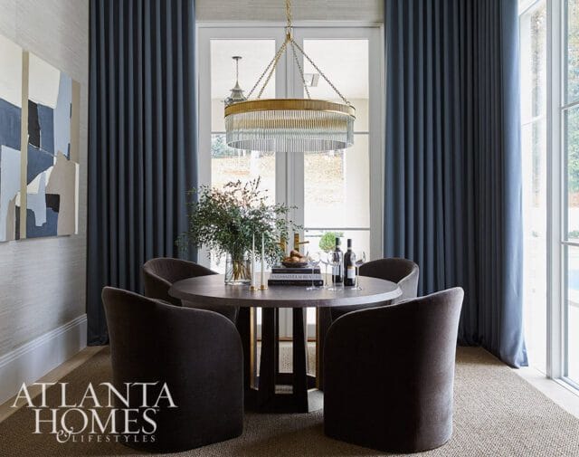 Round dining table with dark upholstered chairs, set with wine and a floral arrangement, beneath a gold and glass chandelier.