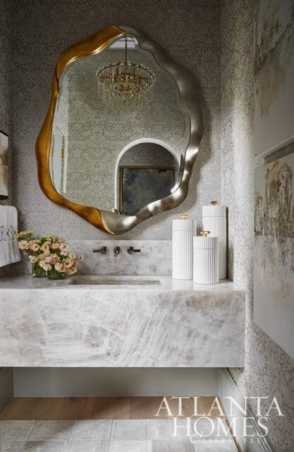 Powder room with a large, organically shaped mirror with gold and silver frame above a marble vanity and sink. Fluted white jars and a floral arrangement sit on the vanity.