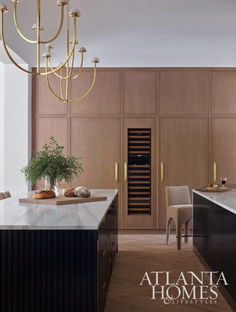 Modern kitchen with dark fluted cabinetry islands topped with marble, a built-in wine fridge, and wood paneled walls. A gold chandelier hangs above.