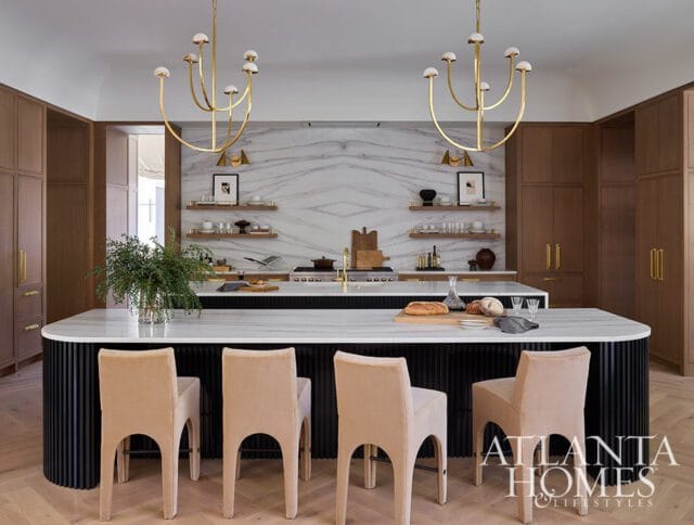 Modern kitchen with a large marble island, fluted black base, and upholstered counter stools. Features wood cabinetry, marble backsplash, and brass chandeliers.