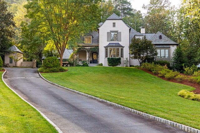 White brick house with gray shutters and a bay window, set on a green lawn with a winding driveway and mature trees.