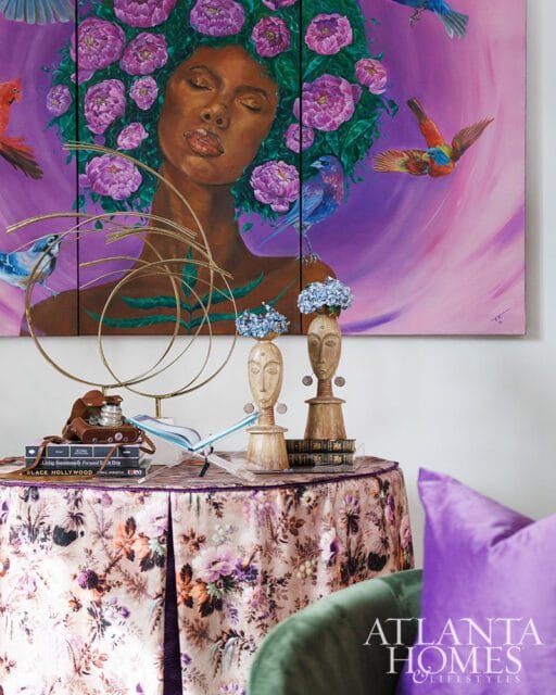 Triptych painting of a woman with flowers in her hair and birds flying around her, displayed above a round table covered with a floral tablecloth.
