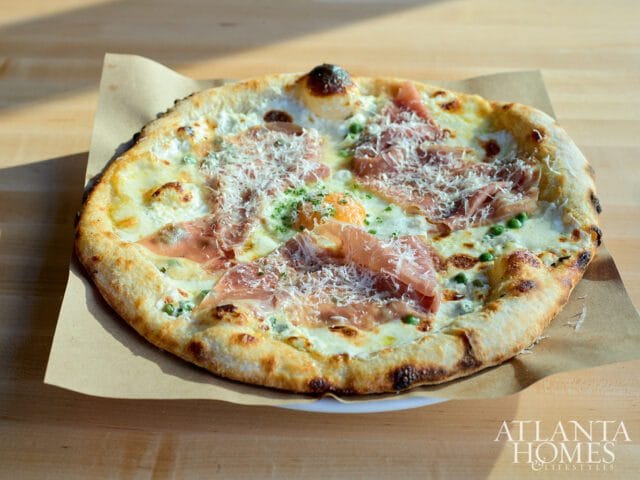 Close-up of a pizza topped with prosciutto, peas, melted cheese, and grated parmesan, served on brown paper over a white plate.