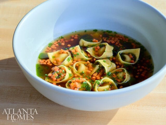 Bowl of tortellini soup with diced carrots and chives in a clear broth, with the Atlanta Homes & Lifestyles logo in the lower left corner.