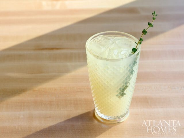 Glass of pale yellow lemonade with ice and a sprig of thyme, set on a light wood surface with diagonal shadows.