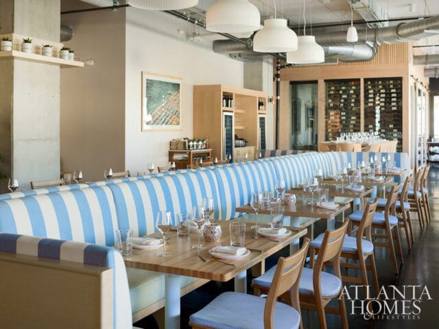 Restaurant dining room with blue and white striped banquettes, light wood tables set with glassware, and a wine display in the background.