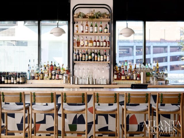 Bar area with wooden stools featuring grey cushions and green straps, set against a backdrop of shelves stocked with liquor bottles and glassware.