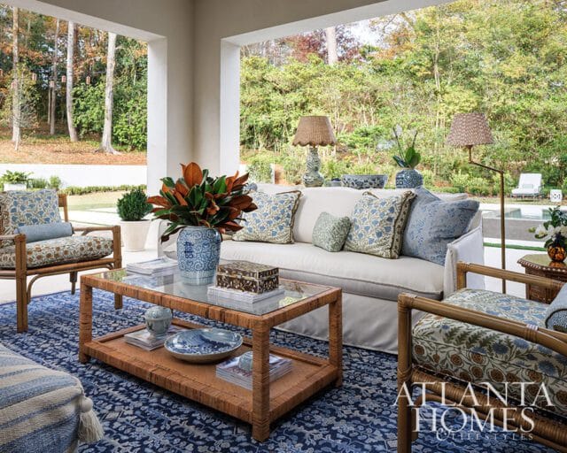 Covered porch with white sofa, rattan coffee table, blue patterned rug, and rattan chairs. Lush green trees and a pool are visible in the background.