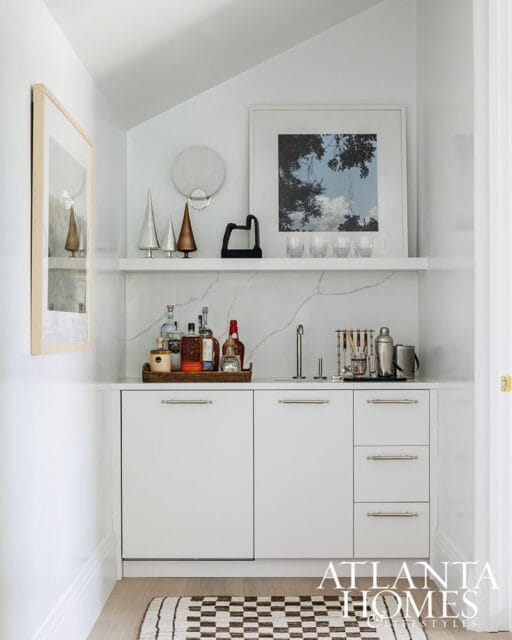White built-in bar with marble backsplash, a tray of liquor bottles, bar tools, and framed artwork on the wall.