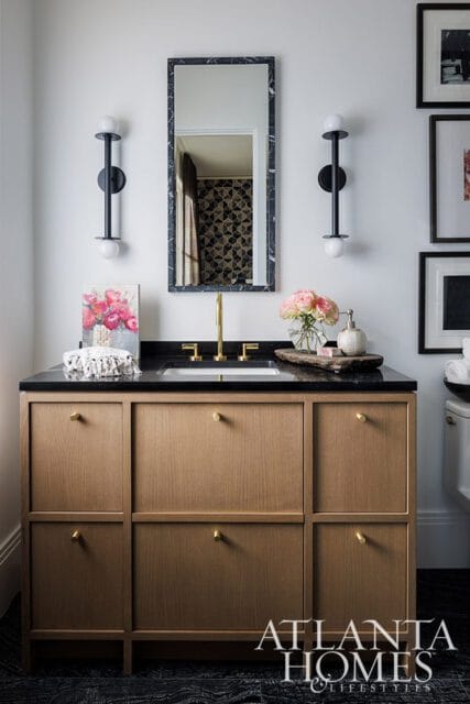Bathroom vanity with light wood cabinetry, black granite countertop, brass faucet, and marble-framed mirror. Modern black sconces flank the mirror.