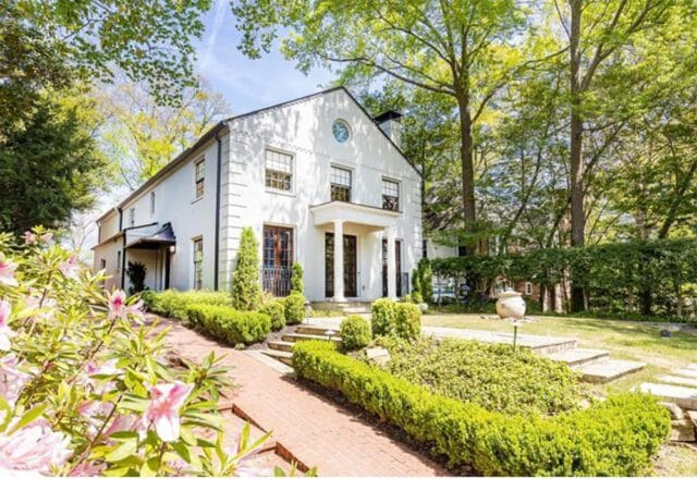 White stucco house with black trim, a covered entryway with columns, and a circular window above the front door. Landscaped garden with brick pathway and manicured hedges.