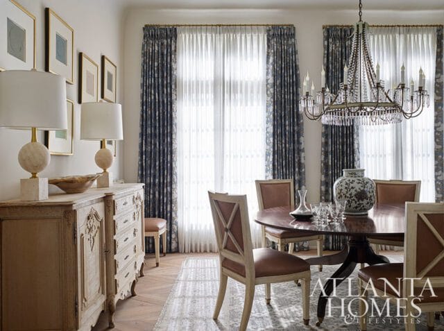 Dining room with a round wooden table, leather upholstered chairs, a vintage dresser with lamps, and patterned curtains framing large windows.