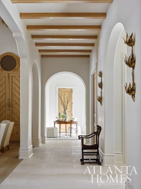 Hallway with arched doorways, exposed wooden ceiling beams, stone tile flooring, and ornate wall sconces. A wooden chair sits near an arched opening.