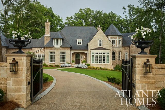 Stone manor with dark slate roof, arched windows, and turrets, accessed by a gravel driveway and ornate black gates.