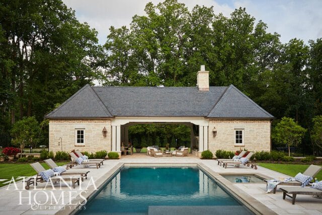 Rectangular swimming pool and hot tub with lounge chairs on a stone patio, in front of a stone pool house with a covered seating area and a slate roof.
