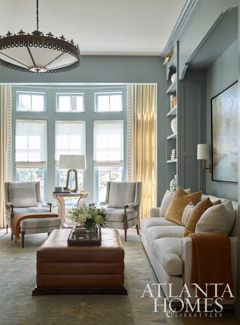 Living room with blue-gray walls, a large bay window with cream blinds, two patterned armchairs, a leather ottoman, and a white sofa with mustard and cream pillows.