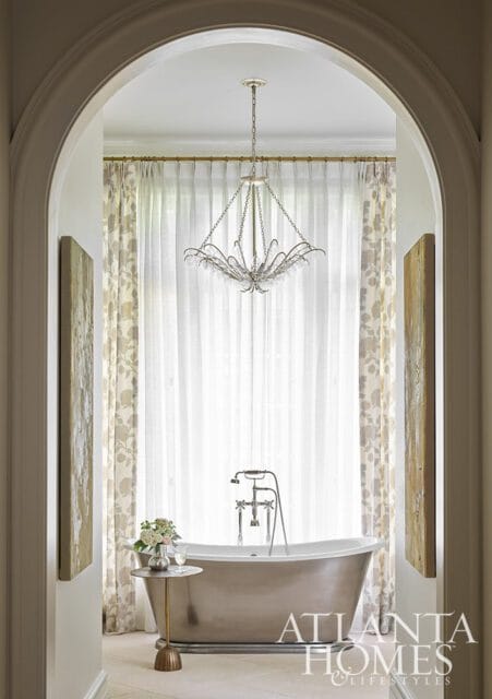 Freestanding bathtub with chrome fixtures in a bathroom with sheer floral curtains and a crystal chandelier.