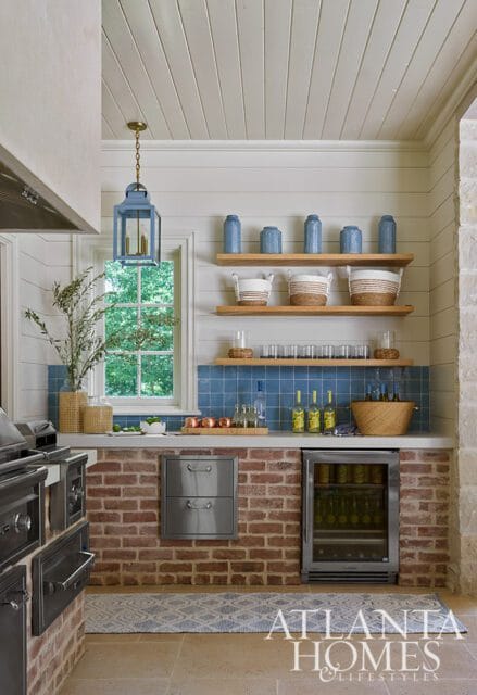 Outdoor kitchen bar with brick base, blue tile backsplash, wooden shelves with blue vases and baskets, and a beverage refrigerator.