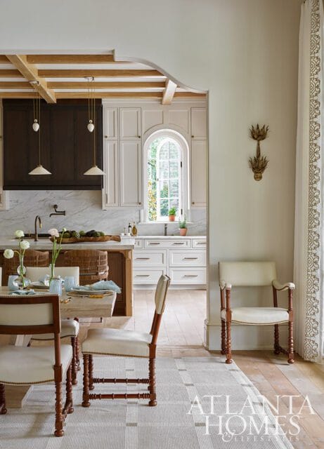 Dining area with light wood table and chairs in foreground, opening to a white kitchen with marble backsplash, dark wood range hood, and arched window.