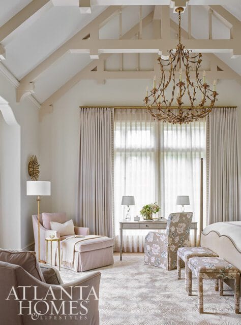 Bedroom with vaulted ceiling featuring exposed beams, a large ornate gold chandelier, and floor-to-ceiling light pink curtains. A chaise lounge, desk with floral chairs, and upholstered benches are visible.