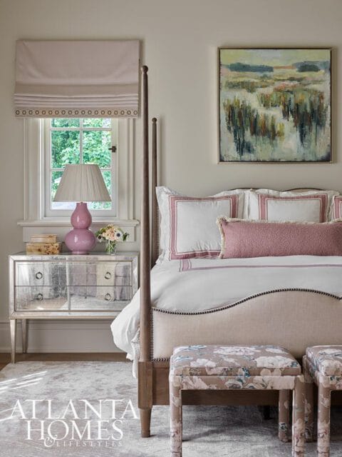 Bedroom with a four-poster bed, mirrored nightstand, pink lamp, and floral upholstered ottomans. A landscape painting hangs above the bed.