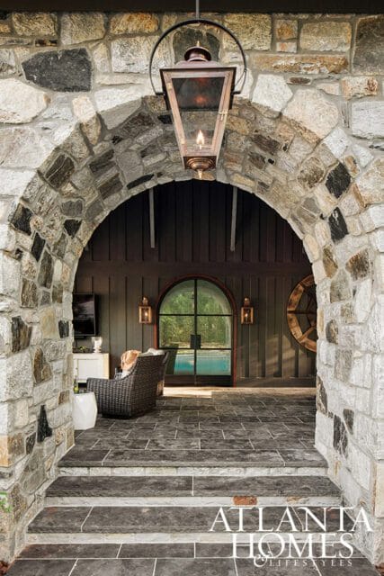 Stone archway entrance with a hanging lantern, leading to a covered patio with dark wood paneling, a large arched window, and a glimpse of a swimming pool.