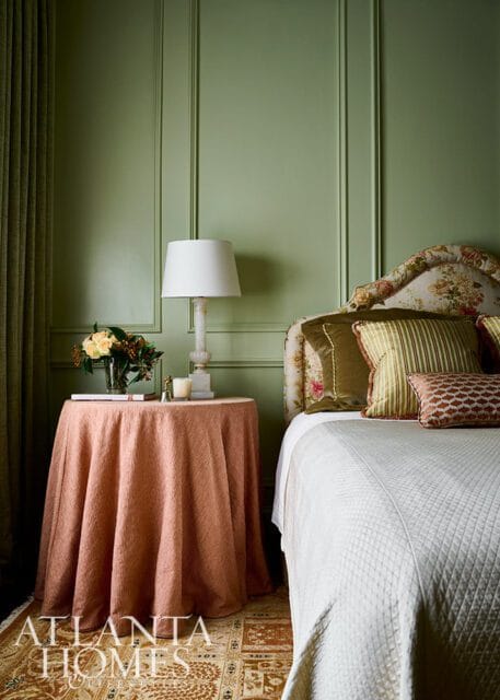 Bedroom with sage green paneled walls, a floral headboard, and a white quilted bedspread. A round side table draped in a coral tablecloth holds a lamp and a vase of flowers.