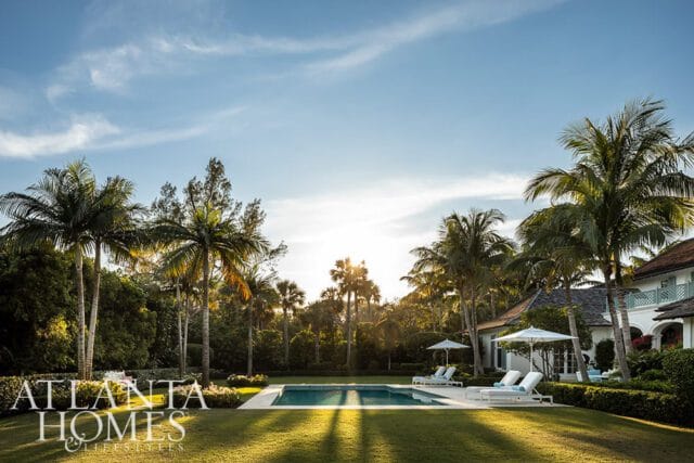 Luxury backyard with a swimming pool, lounge chairs, umbrellas, and palm trees under a bright blue sky with sunbeams.
