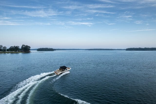 Motorboat speeding across a wide blue lake, leaving a white wake, with tree-lined shores and islands in the distance under a pale blue sky with wispy clouds.