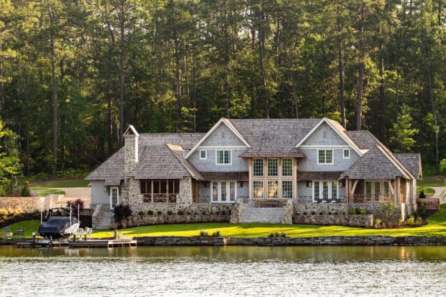 Large stone and shingle lake house with multiple gables, large windows, and a covered boat dock in the foreground.