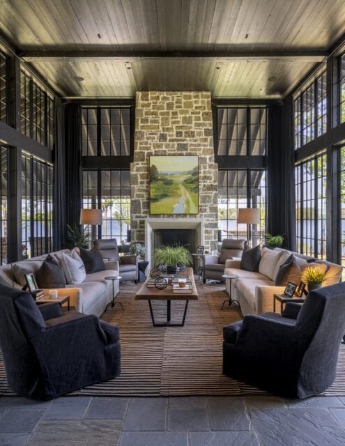 Living room with stone fireplace, large windows overlooking a lake, dark wood ceiling, and seating arranged around a striped rug and wooden coffee table.