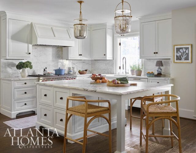 White kitchen with marble countertops and backsplash, custom white cabinetry, a large island with seating, and brass pendant lights.