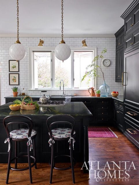 Black kitchen with white subway tile backsplash, dark green granite island, two upholstered bar stools, and brass pendant lights.