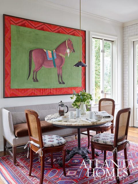 Dining nook with a striped banquette, marble-topped table, cane-backed chairs, and a large painting of a horse above the seating.