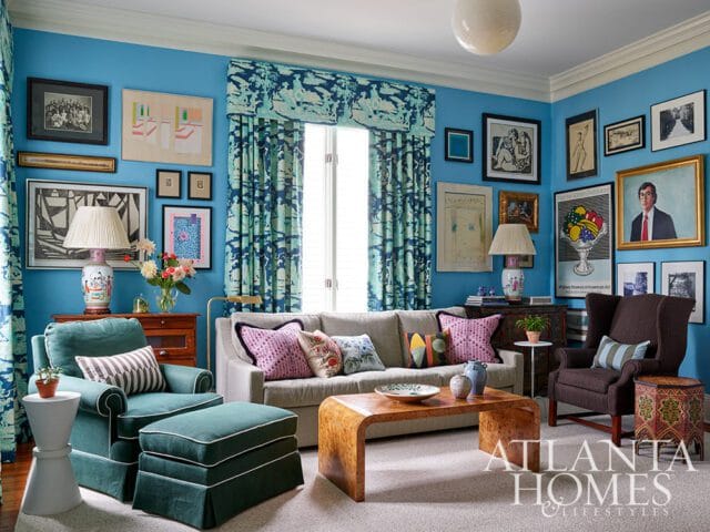 Living room with bright blue walls, teal upholstered chairs, a beige sofa, a burl wood coffee table, and numerous framed artworks.