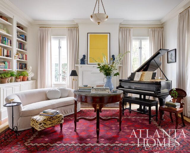 Living room with a grand piano, curved sofa, round table, and bookshelves. A red patterned rug covers the floor and the Atlanta Homes & Lifestyles logo is visible in the lower right corner.