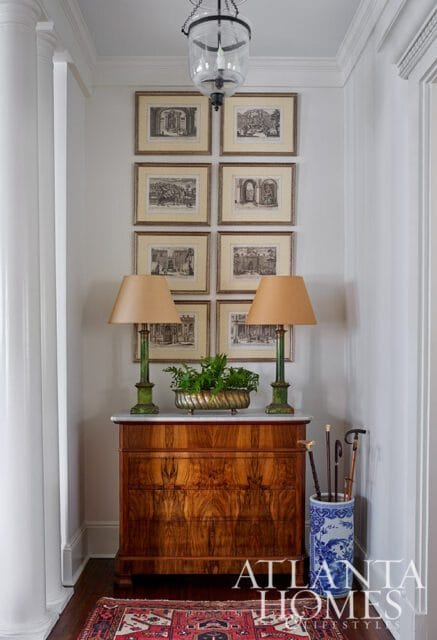 Hallway with a burl wood dresser topped with marble, two green lamps, and a collection of framed architectural prints. A blue and white umbrella stand holds canes.
