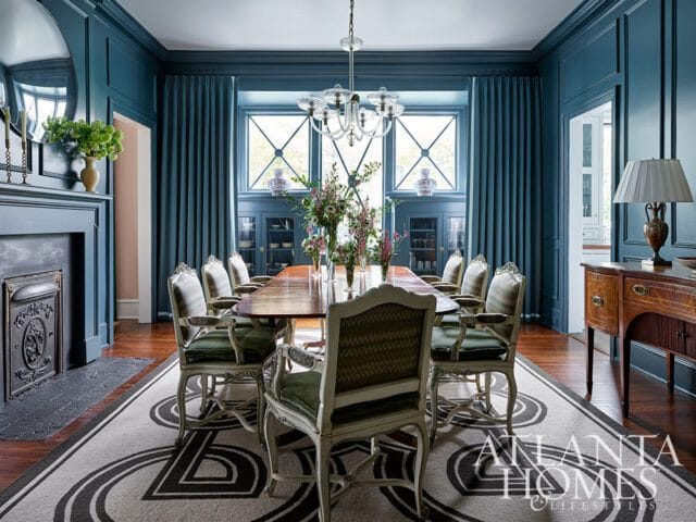 Formal dining room with dark teal walls, a large wooden table set with ten chairs, a fireplace, and a patterned rug.