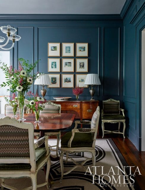 Dining room with dark blue walls, a wooden dining table, upholstered chairs, a buffet, and a gallery wall of nine framed prints.