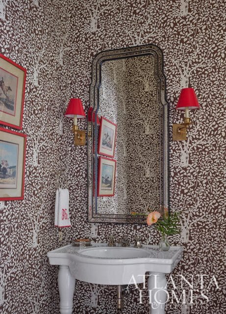 Powder room with brown and white tree-patterned wallpaper, a white pedestal sink, a decorative mirror, red lampshades, and framed artwork.