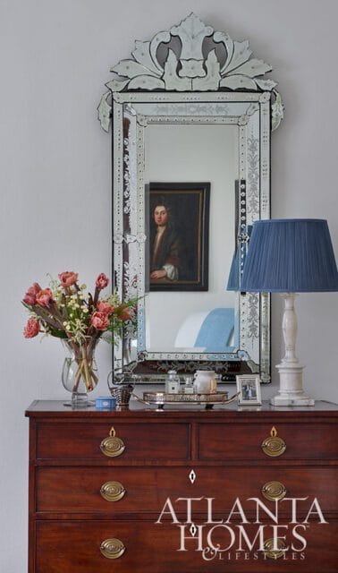 Dark wood dresser with brass hardware topped by a large ornate mirror reflecting a portrait, a vase of pink flowers, and a blue lampshade.